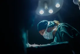 Surgeon wearing a mask and cap, focused intently under bright surgical lights in an operating room.