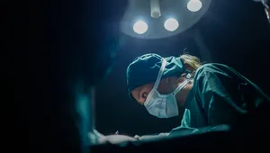Surgeon wearing a mask and cap, focused intently under bright surgical lights in an operating room.