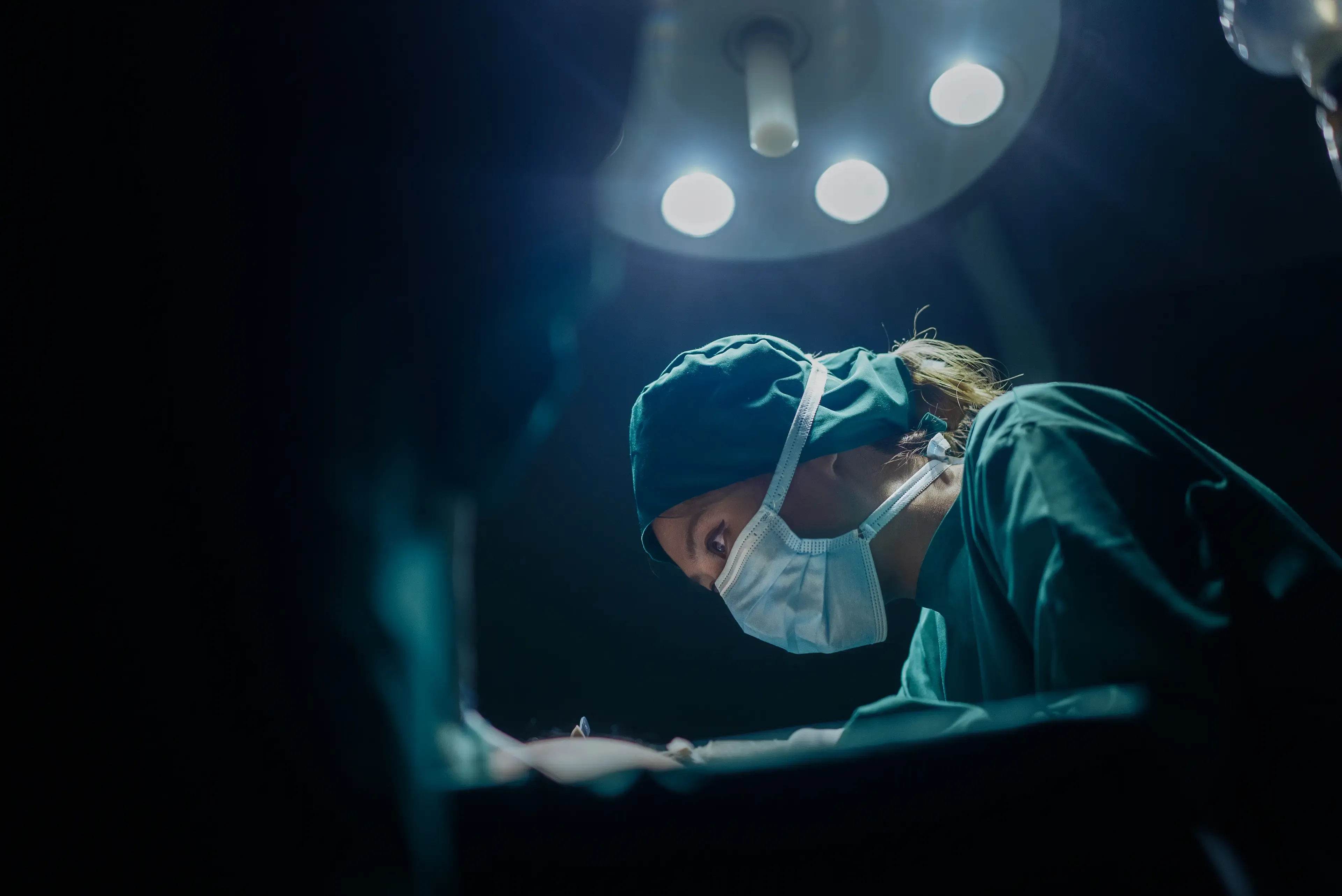 Surgeon wearing a mask and cap, focused intently under bright surgical lights in an operating room.