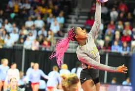 Volleyball player with pink braids jumps to spike the ball during a match, with a focused crowd in the background.