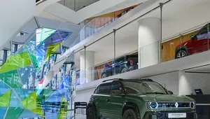 A green SUV is displayed inside a modern showroom with glass and colorful geometric accents, alongside a mirrored ceiling.
