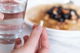Hand holding a white capsule next to a glass of water, with a plate of food in the background.