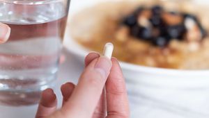 Hand holding a white capsule next to a glass of water, with a plate of food in the background.