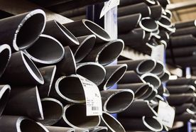 Stacks of dark metal pipes with labels, stored in a warehouse, partially lit by overhead lighting.