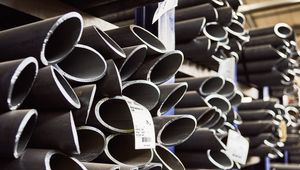 Stacks of dark metal pipes with labels, stored in a warehouse, partially lit by overhead lighting.