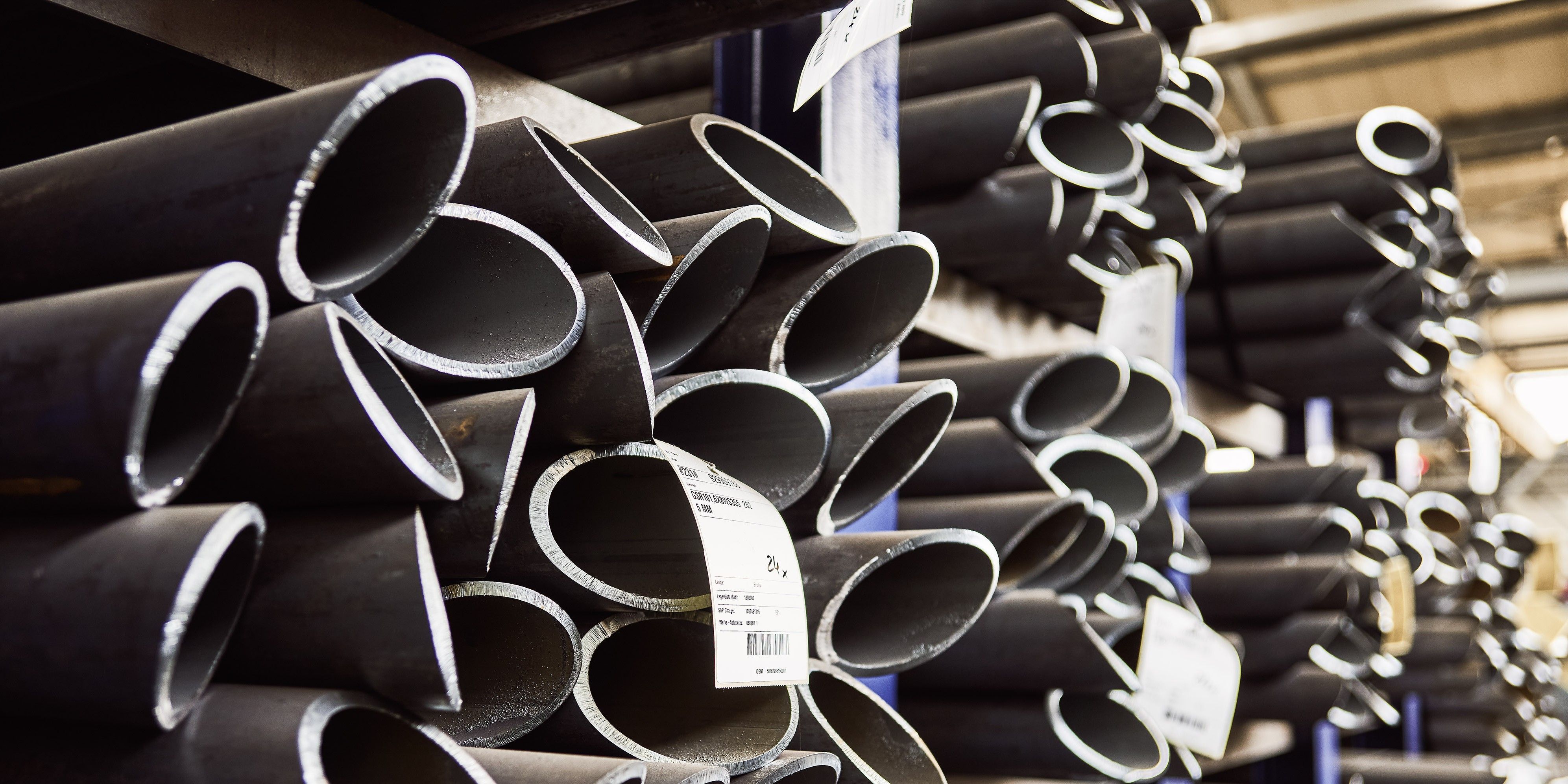 Stacks of dark metal pipes with labels, stored in a warehouse, partially lit by overhead lighting.