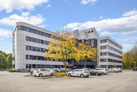 A modern office building with large windows, surrounded by parking spaces and trees with autumn foliage under a blue sky.