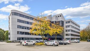 A modern office building with large windows, surrounded by parking spaces and trees with autumn foliage under a blue sky.