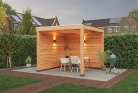 Wooden gazebo on a patio with a table, chairs, and a grill. Surrounded by a hedge, under a cloudy sky with neighboring houses in the background.