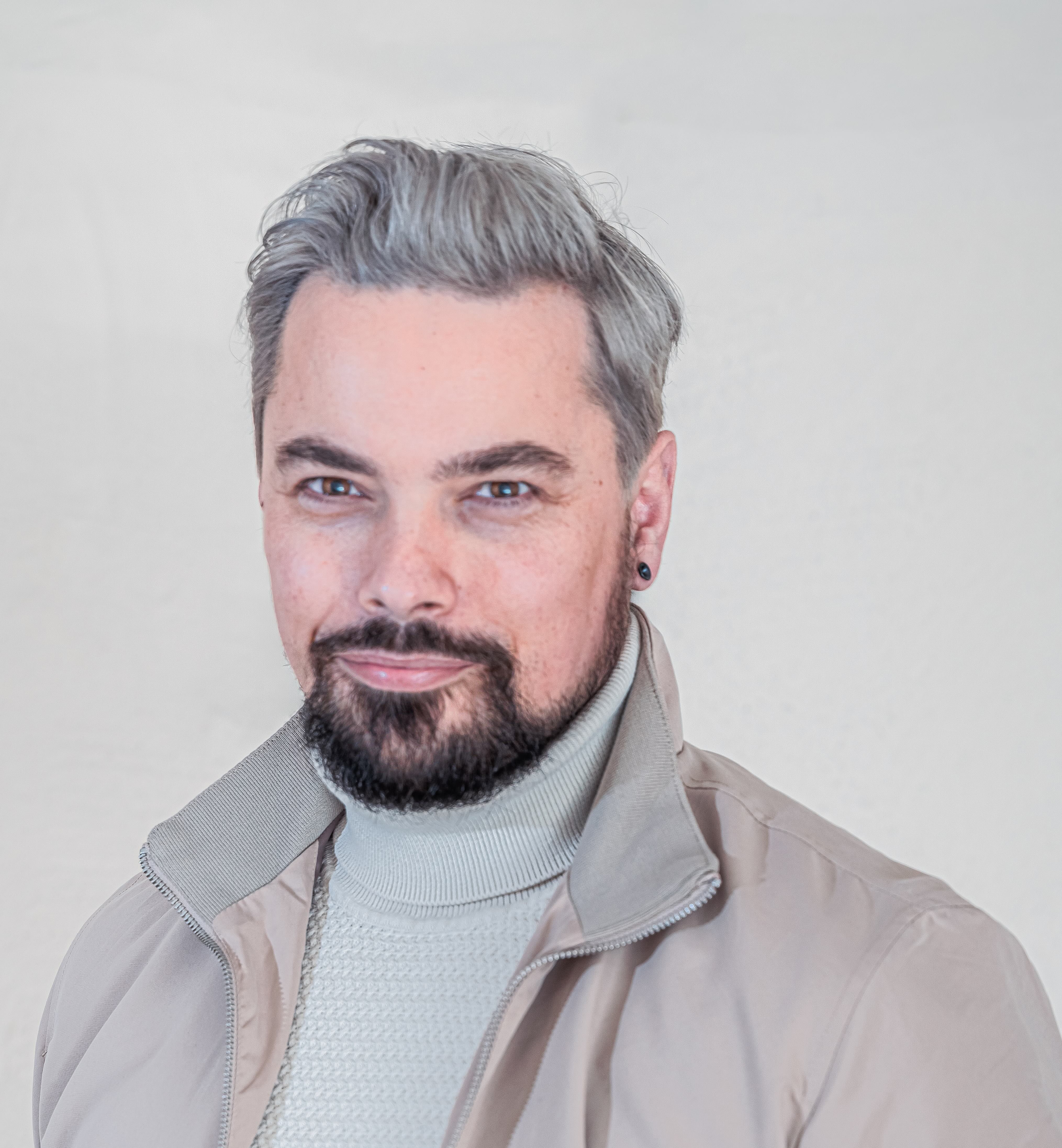 A man with gray hair and a beard, wearing a beige jacket and a light sweater, against a plain background.