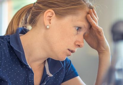 A woman with a ponytail looks stressed, resting her forehead on her hand. She wears a blue collared shirt with small white dots.