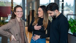 Three people in a modern office setting are smiling and chatting, with plants and windows in the background.