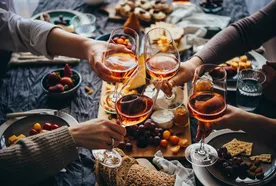 Four people toast with glasses of rosé wine over a table set with assorted snacks, grapes, bread, and cheese.