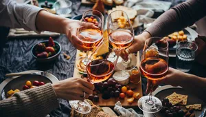 Four people toast with glasses of rosé wine over a table set with assorted snacks, grapes, bread, and cheese.