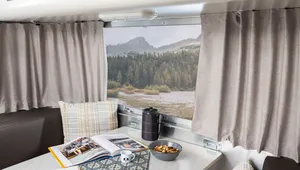 Alt text: "Interior of an RV or camper trailer showing a dinette table with an open magazine, notebook and pen, a small mug, a bowl of trail mix, and a thermos, framed by curtained windows revealing a scenic mountain and forest lake view outside.
