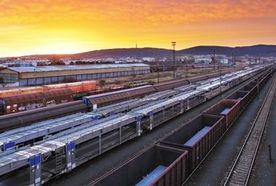 Train station at sunset with multiple tracks and trains, industrial buildings in the background, under a vibrant orange and purple sky.
