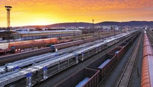 Train station at sunset with multiple tracks and trains, industrial buildings in the background, under a vibrant orange and purple sky.