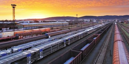 Train station at sunset with multiple tracks and trains, industrial buildings in the background, under a vibrant orange and purple sky.