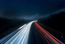 A highway at night with long-exposure light trails; white on the left and red on the right, surrounded by dark, tree-lined landscape.