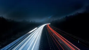 A highway at night with long-exposure light trails; white on the left and red on the right, surrounded by dark, tree-lined landscape.