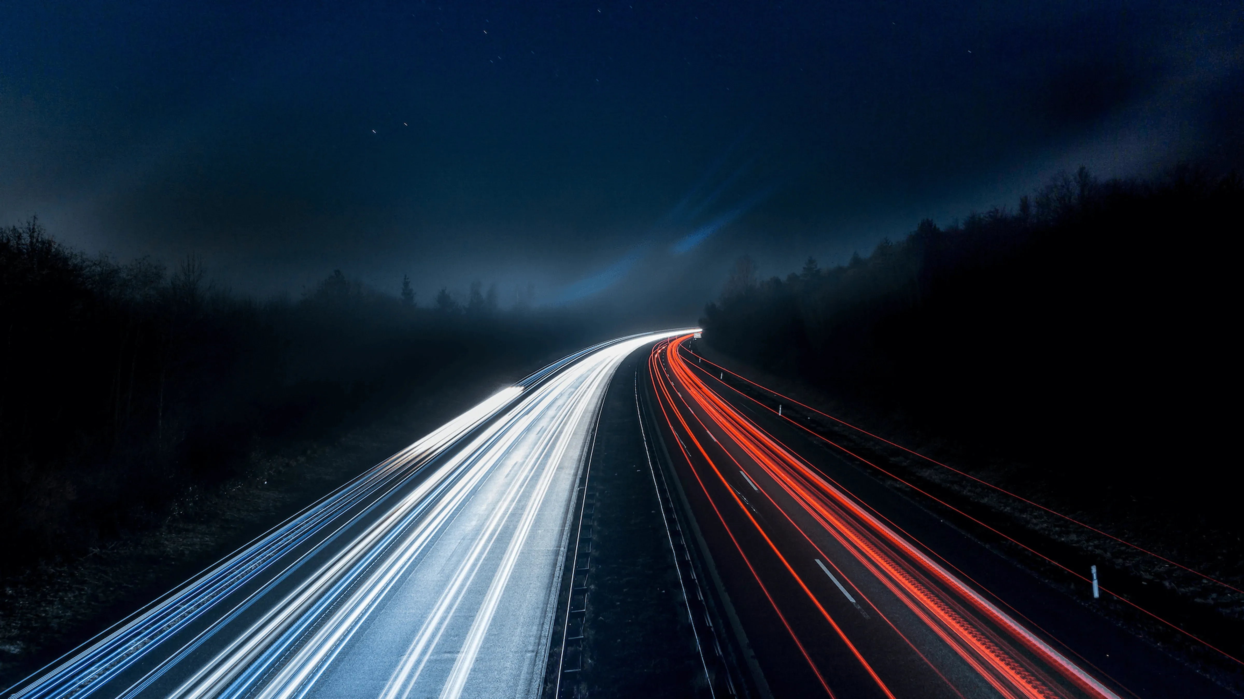 A highway at night with long-exposure light trails; white on the left and red on the right, surrounded by dark, tree-lined landscape.
