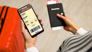 A person holding a smartphone displaying a digital train ticket from Paris to Milan, while holding a passport and boarding passes, with a red suitcase in the background.