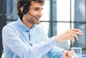 Man in a striped shirt wearing a headset, sitting at a glass table in a bright office, gesturing while speaking.