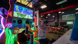 Person playing a neon-lit arcade game featuring a SpongeBob theme in a dimly lit leisure center. Golf is shown on a TV in the background.