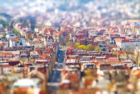 Tilt-shift photo of a European cityscape with red-roofed buildings, creating a miniature effect. Central road flanked by colorful architecture.