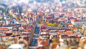 Tilt-shift photo of a European cityscape with red-roofed buildings, creating a miniature effect. Central road flanked by colorful architecture.