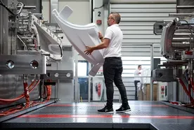 A worker in a factory operates a large machine, handling a curved, white plastic component in an industrial setting.