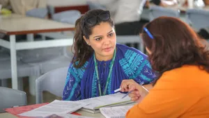 Two women sitting at a table, engaged in a discussion, with documents and notebooks in front of them in a classroom setting.