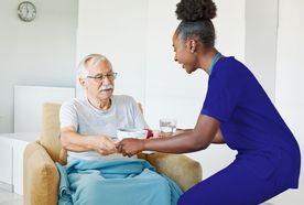 A caregiver in blue scrubs hands a cup to an elderly man wrapped in a blue blanket, seated in a comfortable chair.