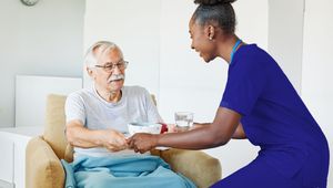 A caregiver in blue scrubs hands a cup to an elderly man wrapped in a blue blanket, seated in a comfortable chair.