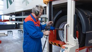 A mechanic in blue coveralls uses a power tool to work on a truck wheel in a garage.