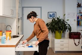 A healthcare professional in a brown sweater looks through a medical supply drawer in a clinical room, with a sharps disposal bin and hand sanitiser visible on the counter.