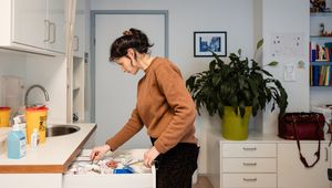 A healthcare professional in a brown sweater looks through a medical supply drawer in a clinical room, with a sharps disposal bin and hand sanitiser visible on the counter.