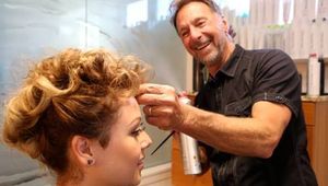 A hairstylist smiles as he sprays a woman's styled curly hair in a salon.