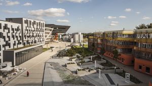 Modern campus with geometric buildings, including a black and white structure and an orange one, surrounded by pathways and greenery.