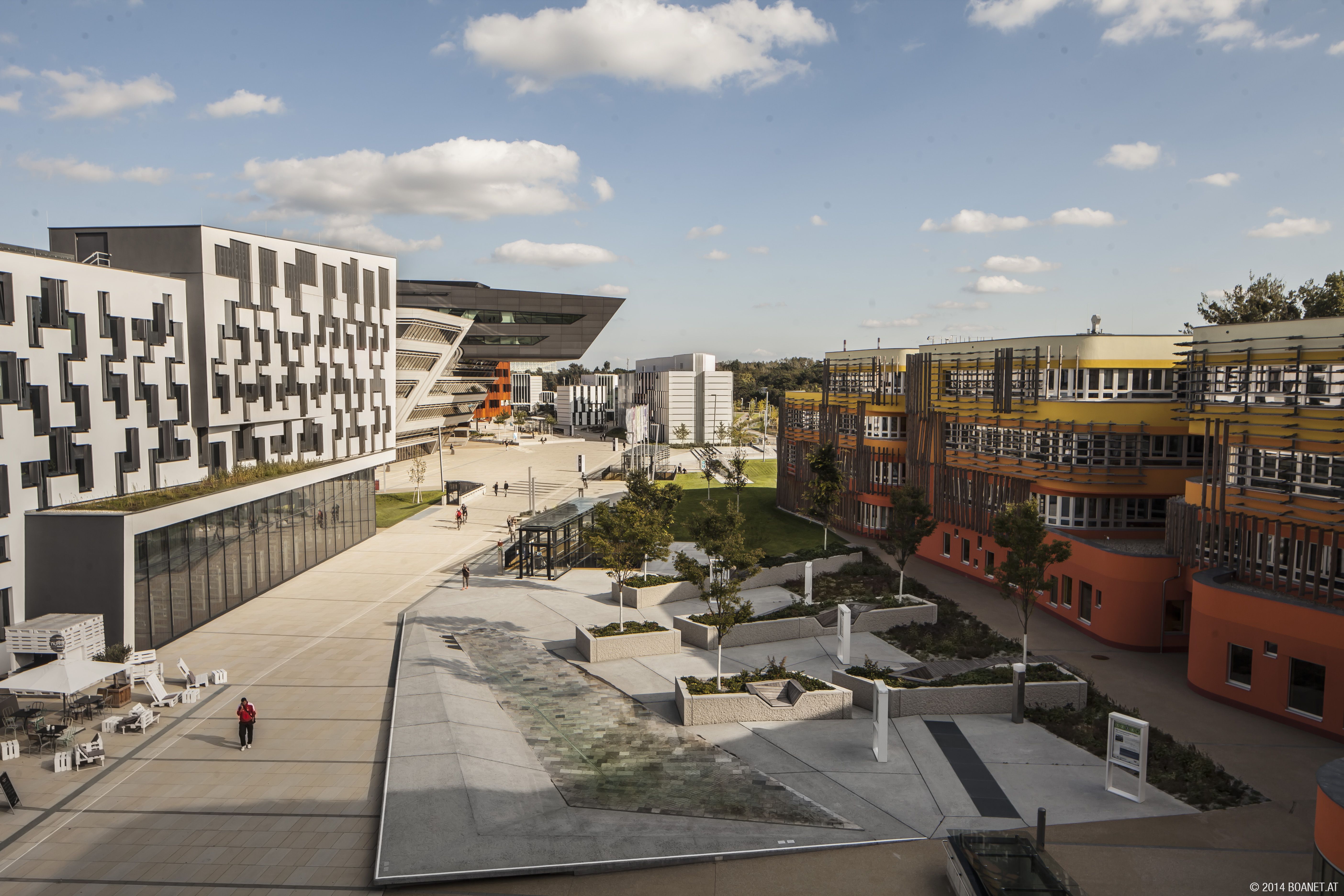 Modern campus with geometric buildings, including a black and white structure and an orange one, surrounded by pathways and greenery.