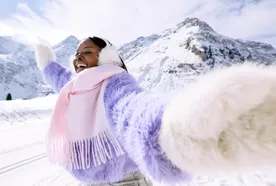Person in a purple sweater and earmuffs joyfully extends arms in a snowy mountain landscape, with a pink scarf and bright sky overhead.
