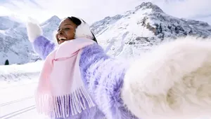 Person in a purple sweater and earmuffs joyfully extends arms in a snowy mountain landscape, with a pink scarf and bright sky overhead.