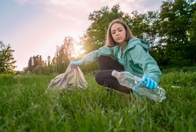 A young woman in a green jacket picks up a plastic bottle in a park, placing it into a bag, with the sun setting behind her.