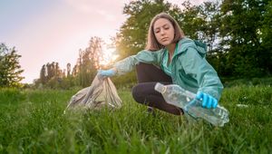 A young woman in a green jacket picks up a plastic bottle in a park, placing it into a bag, with the sun setting behind her.