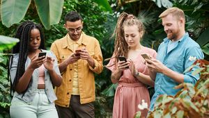 Four people stand together in a lush green setting, all focused on their smartphones.