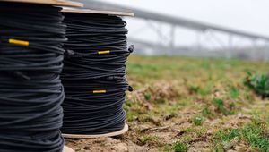 Spools of black cables with yellow labels on grass, and a blurred structure in the background under a cloudy sky.