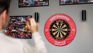 Person aiming a dart at a dartboard on a wall with photos. Dartboard is branded with "Dartshopper" and "Winmau."