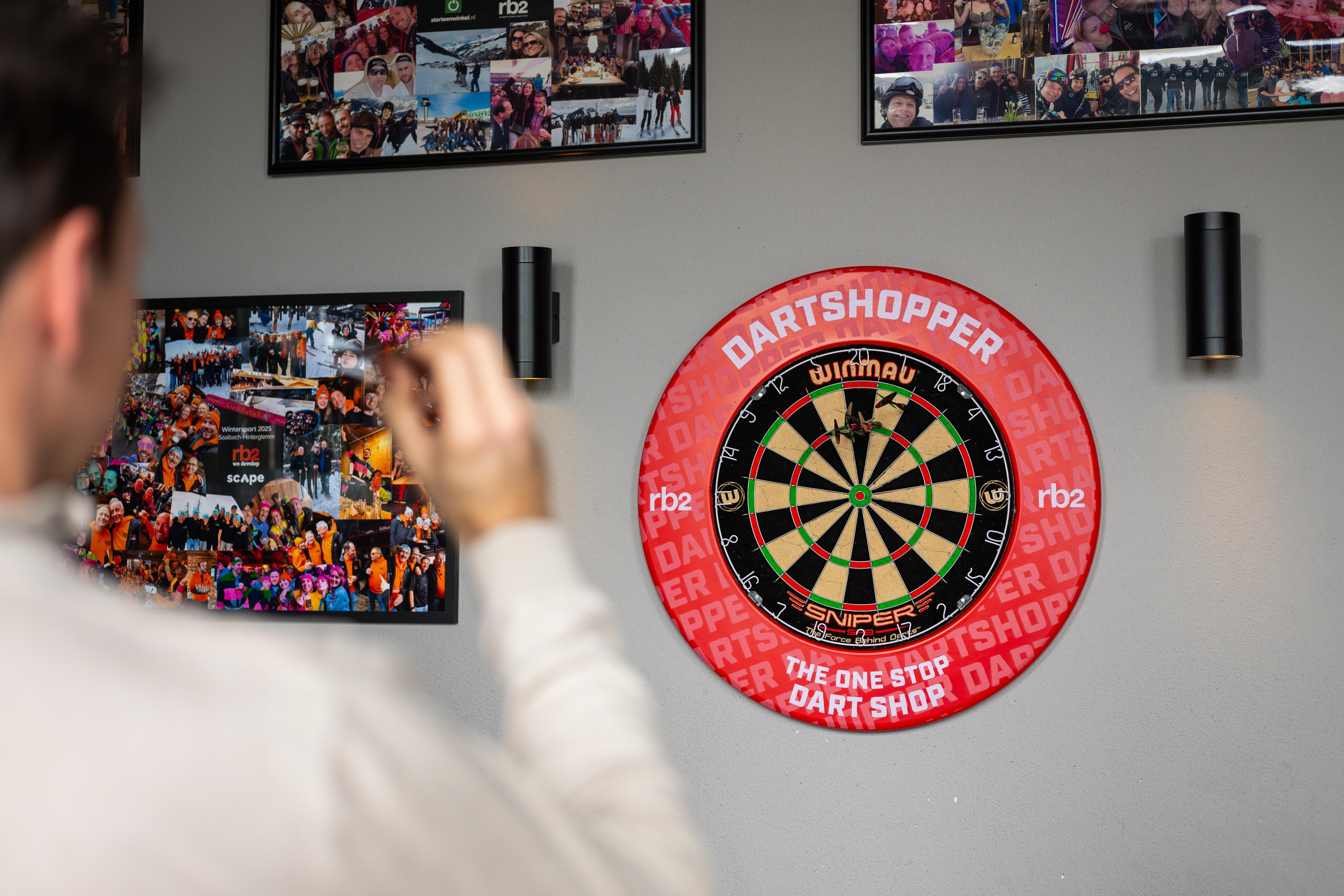 Person aiming a dart at a dartboard on a wall with photos. Dartboard is branded with "Dartshopper" and "Winmau."