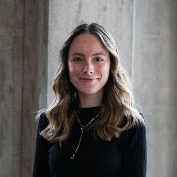 Smiling woman with wavy hair wearing a black top and gold necklace, standing against a concrete wall background.