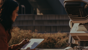 A woman with a notepad sits on the ground, interacting with a humanoid robot outdoors.